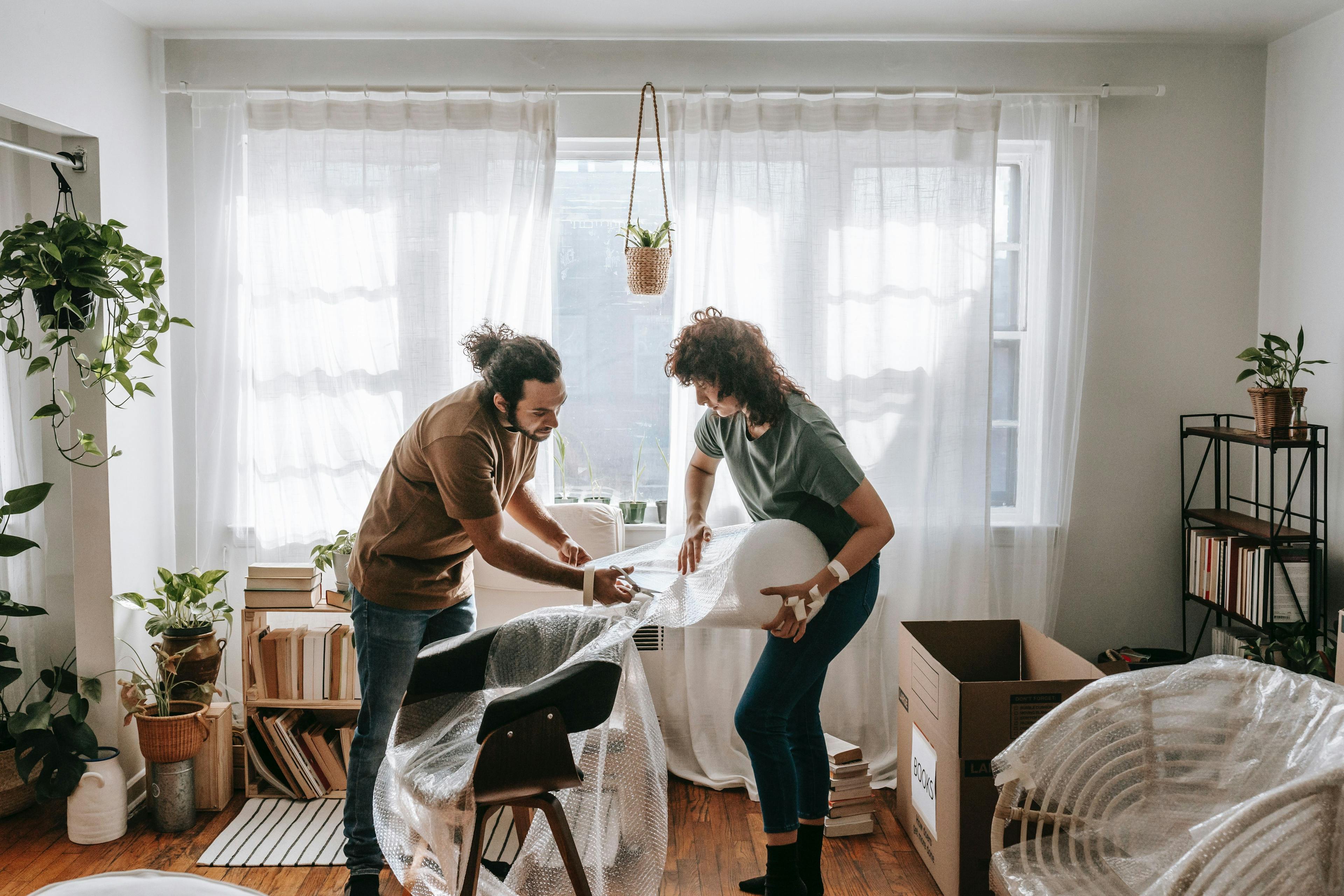 Two persons wrapping a chair in bubble wrap in a cosy messy room.
