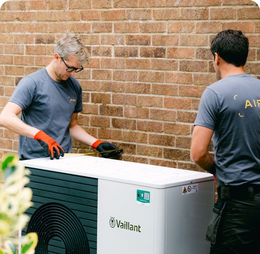 Two heat pump installers from Aira placing a heat pump next to a brick wall