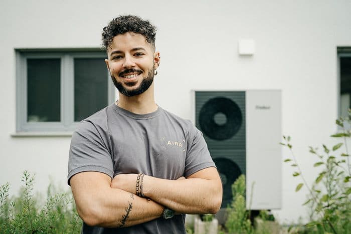 Techniker steht vor einem Haus mit verschränkten Armen, im Hintergrund steht eine Wärmpumpe