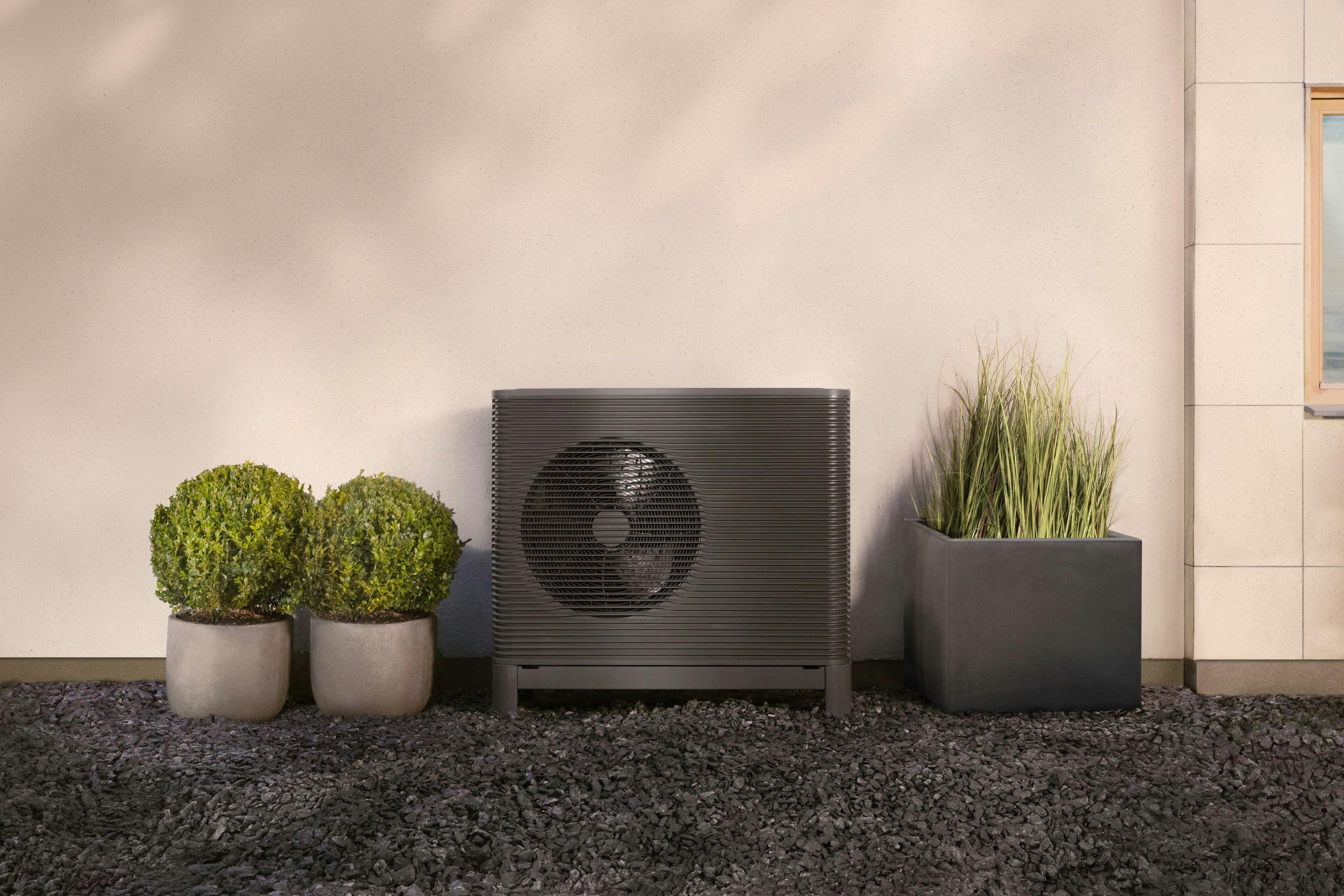 Aira heat pump outdoor unit face on against a wall with some potted plants either side