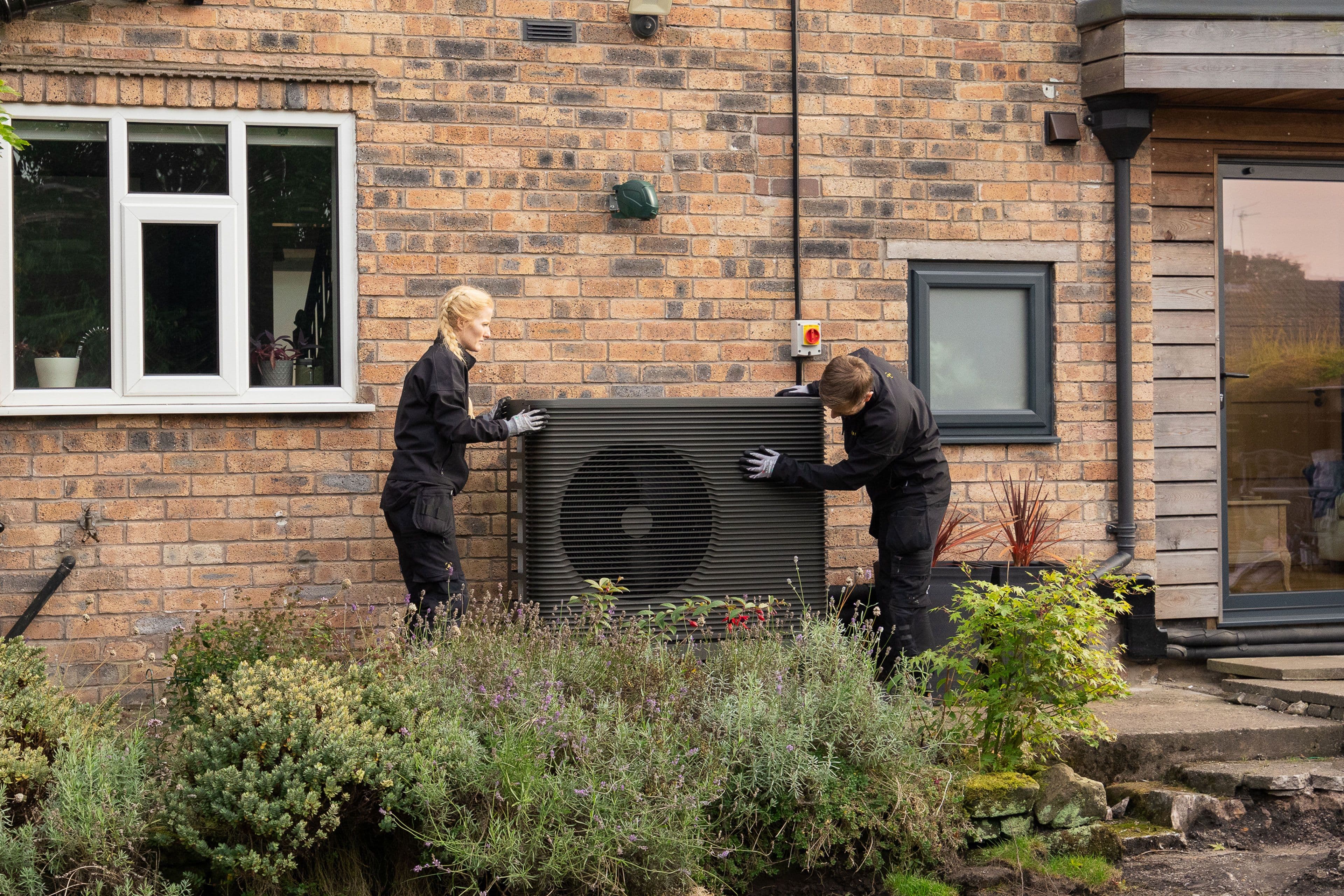 Zwei Installateure von Aira installieren ein Außengerät eines Wärmepumpen-Systems bei einem Kunden vorm Haus