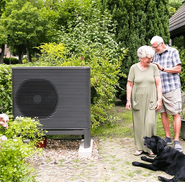 älteres Ehepaar steht mit Hund neben Wärmepumpen Außeneinheit im Garten und lacht