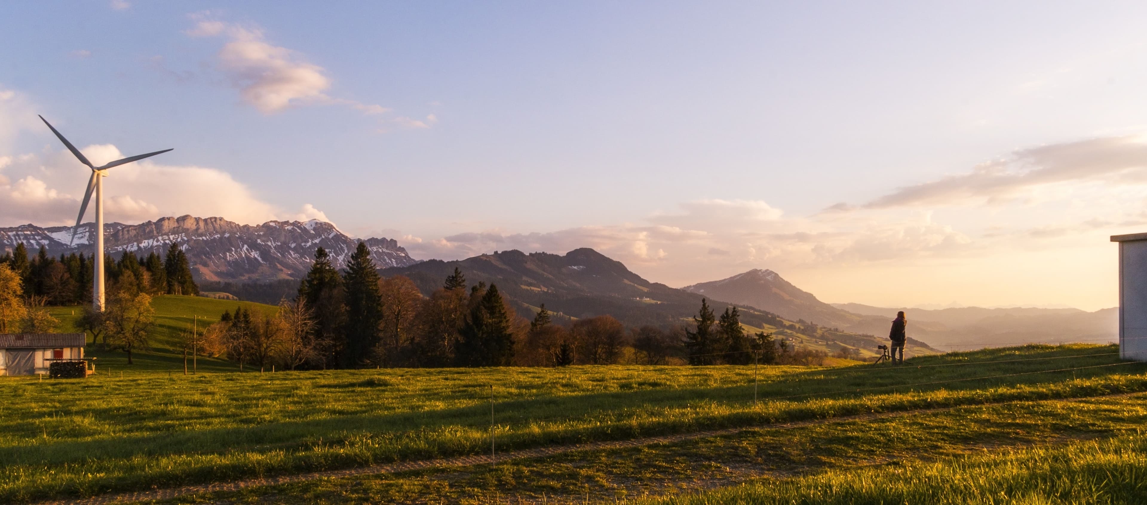 A lone windmill standing on a field with mighty mountains in the background. A person is standing looking at it in the forefround.