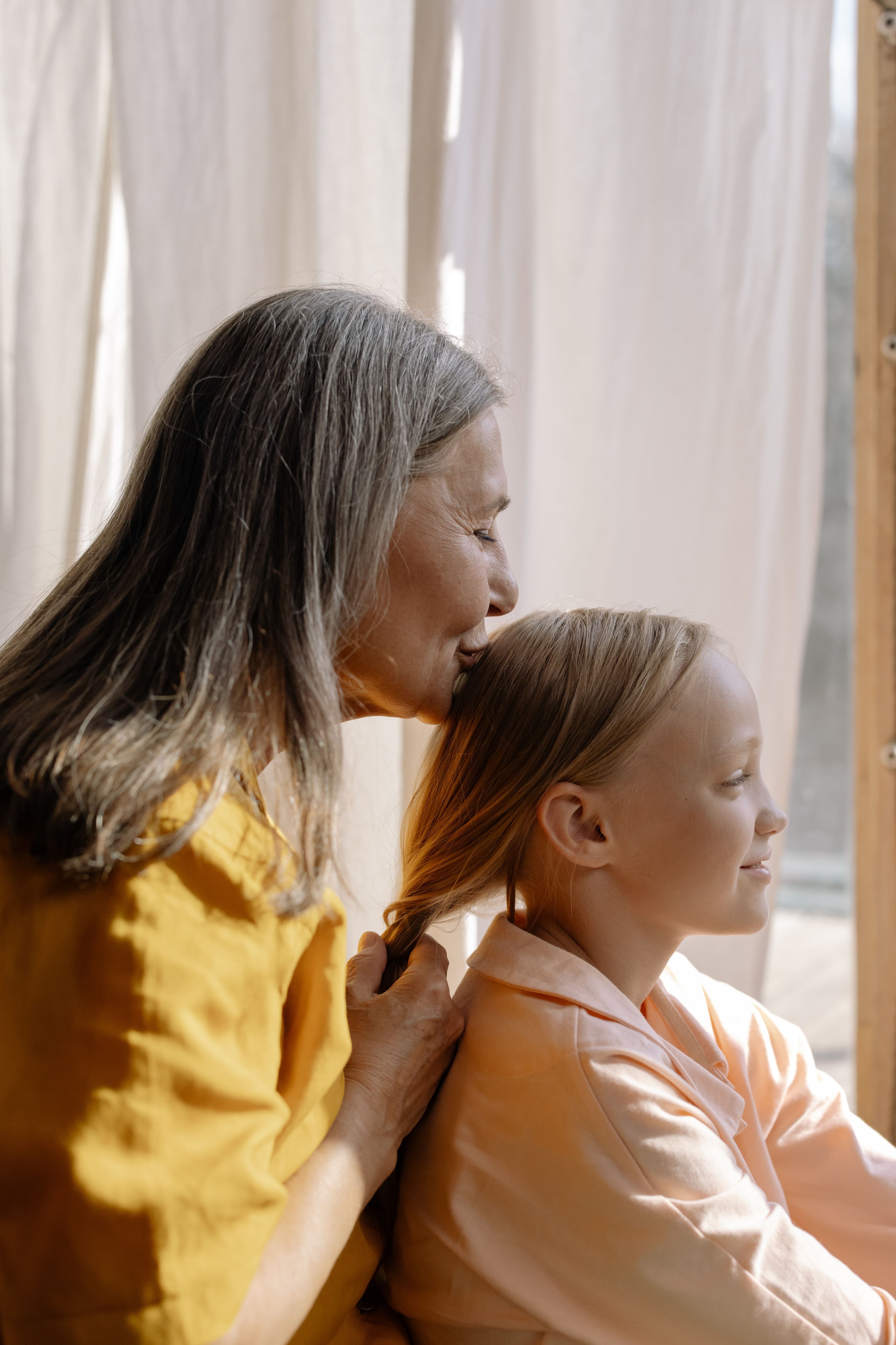 Elderly woman braiding a young girl's hair and kissing her on the head.