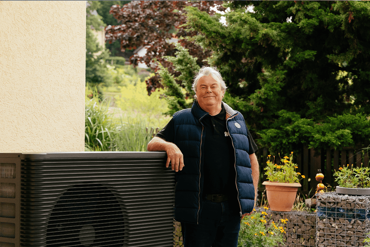 Eine zufriedene Person steht neben seiner Wärmepumpe im Garten vor der Hauswand
